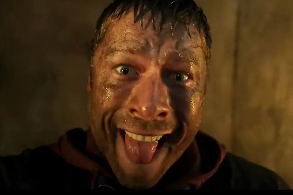 Glenn Powell, lead actor of the film The Running Man, seen in a promotional photo from the film. He is dirty and sweaty with his tongue sticking out and a crazed expression.