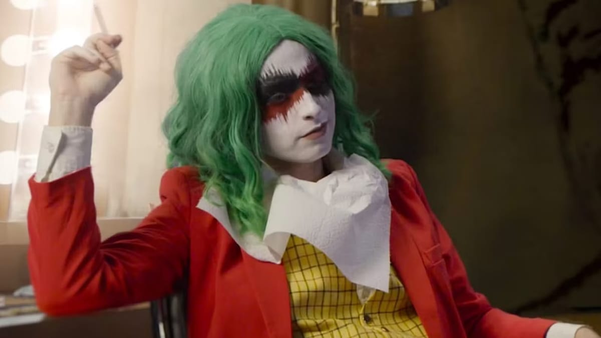 An image of The People's Joker lead actress Vera Drew, in a red suit jacket with yellow checkered shirt and bold white and red makeup, in a shot parodying the 2019 film Joker.