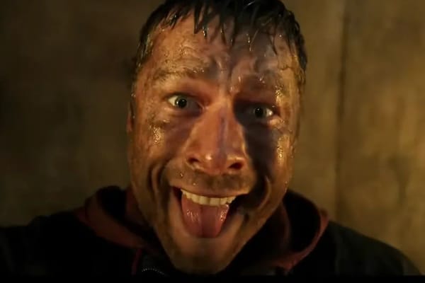 Glenn Powell, lead actor of the film The Running Man, seen in a promotional photo from the film. He is dirty and sweaty with his tongue sticking out and a crazed expression.